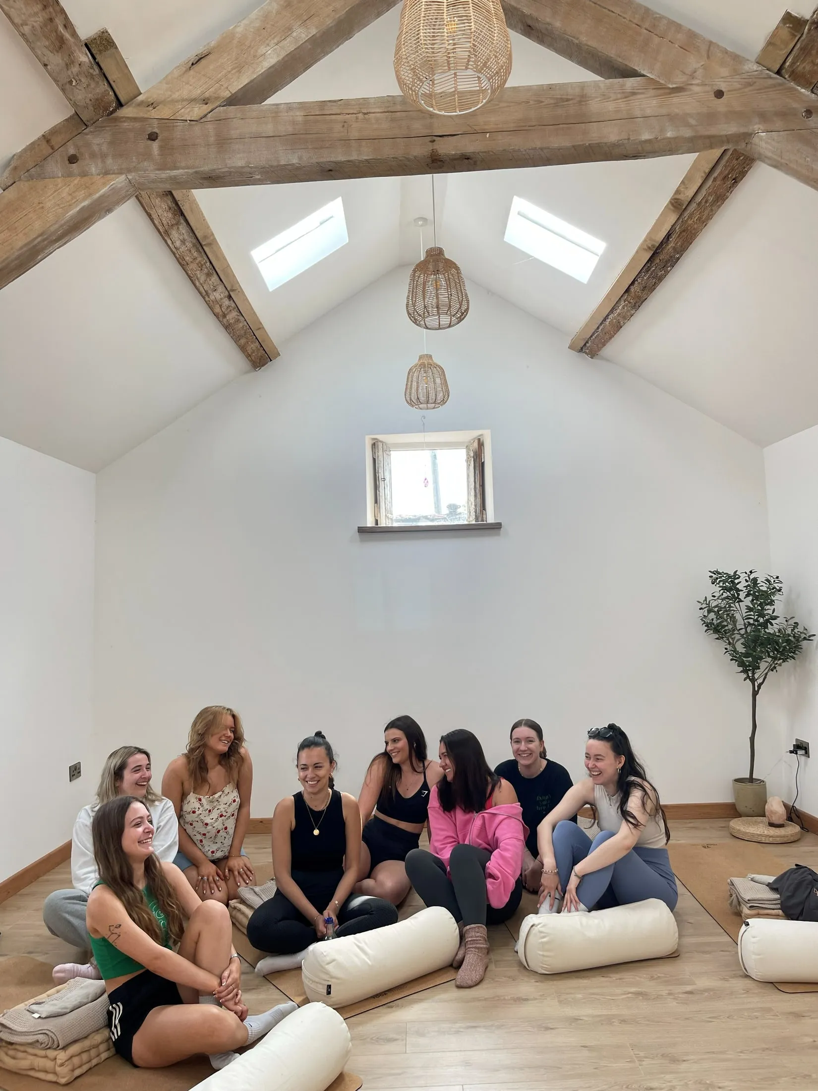 A group of women smiling and laughing on the floor of a white studio under wooden beams