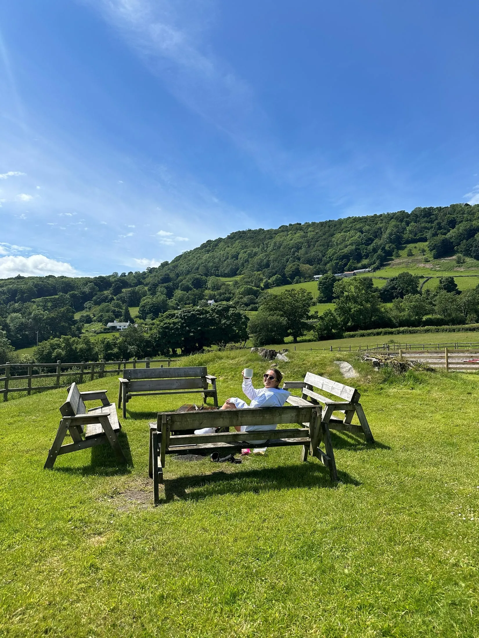 A woman in a white dressing gown sitting on a bench and raising her mug, green hills and blue sky behind