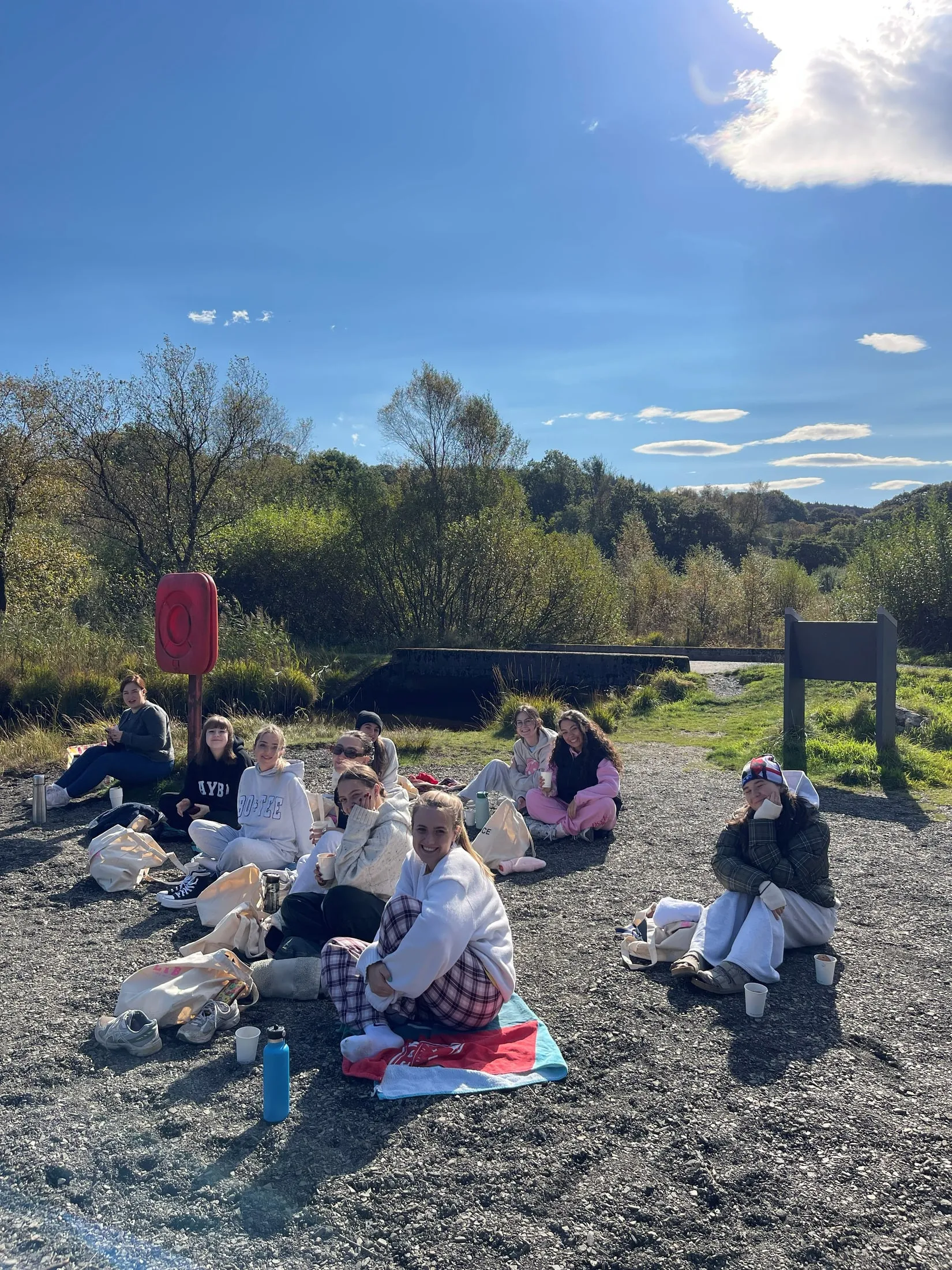 A group of women in loungewear sitting on gravel in the sun with paper cups, trees behind