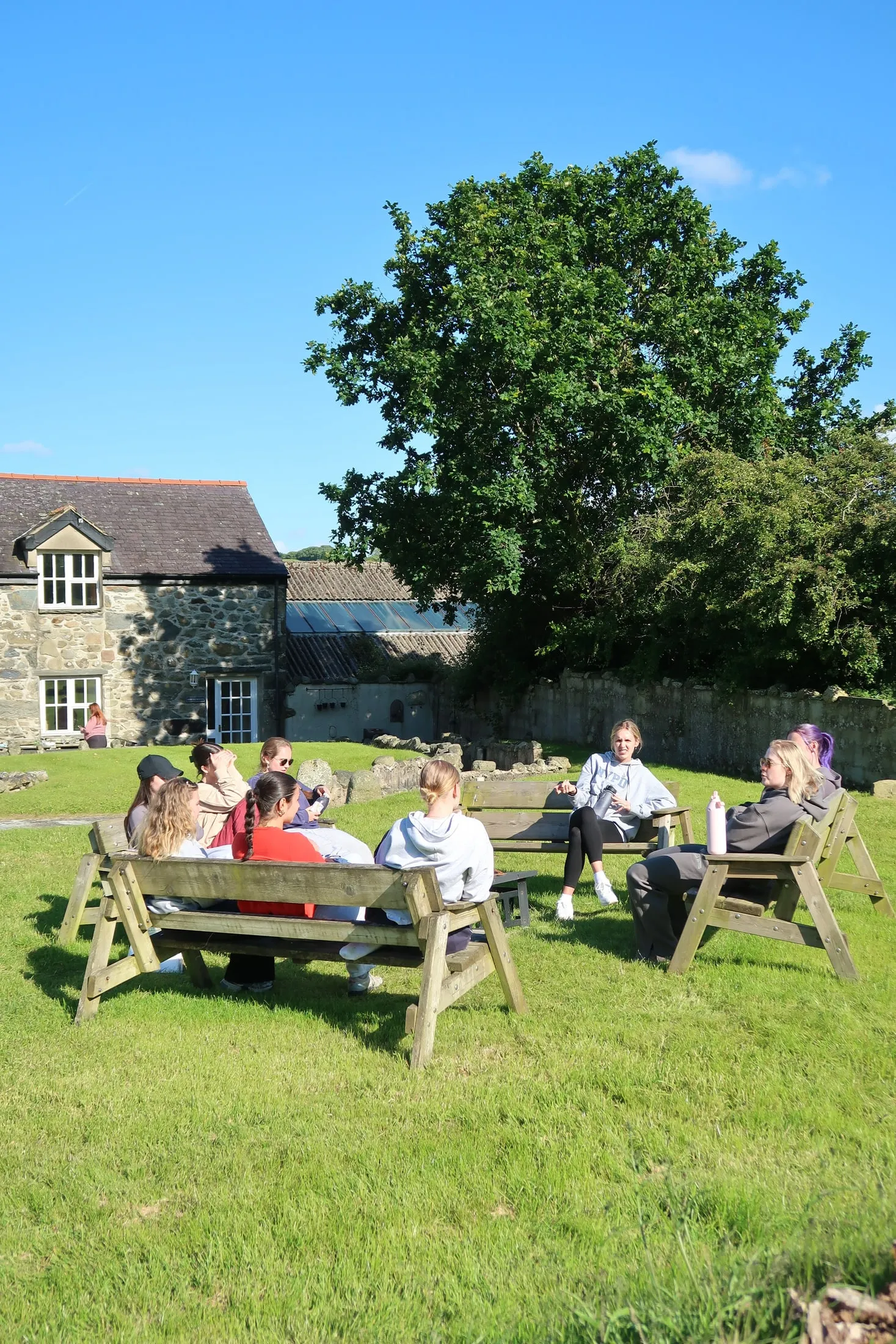 Women sitting on a circle of wooden benches on a sunny lawn, a stone house and a big tree behind