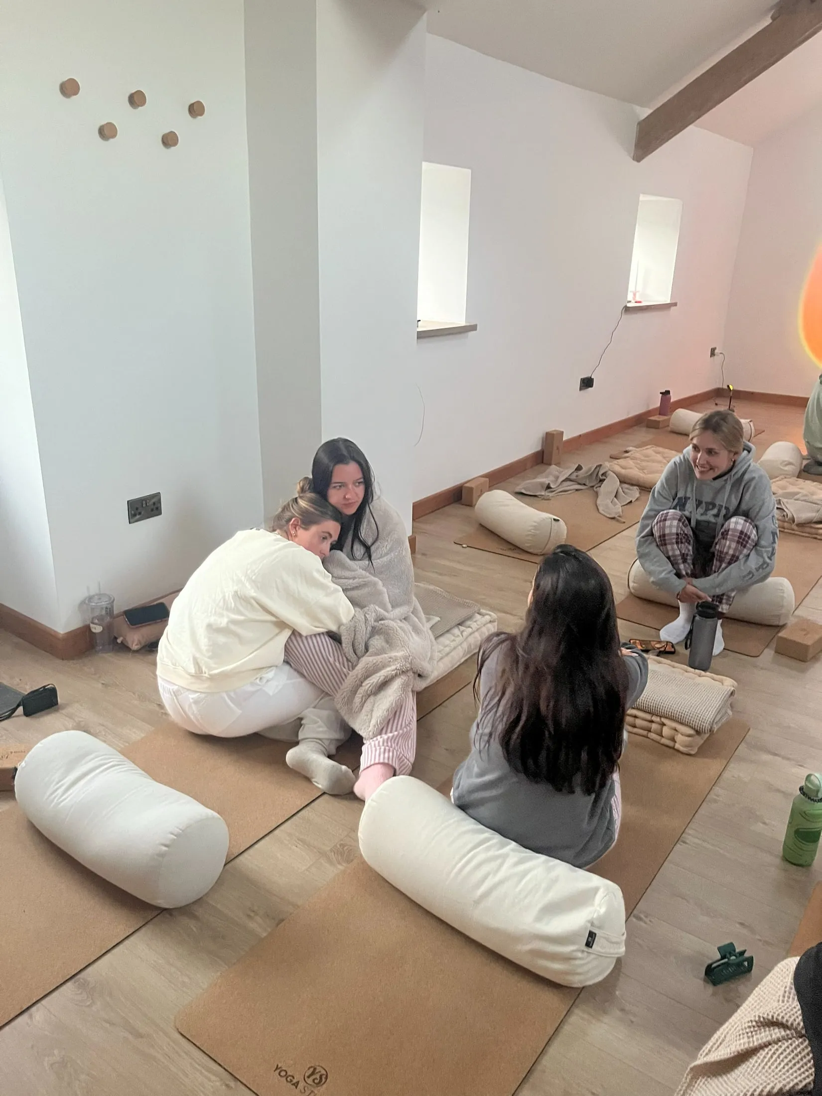 Two women cuddled up together on cork yoga mats in a bright white studio, others sitting nearby