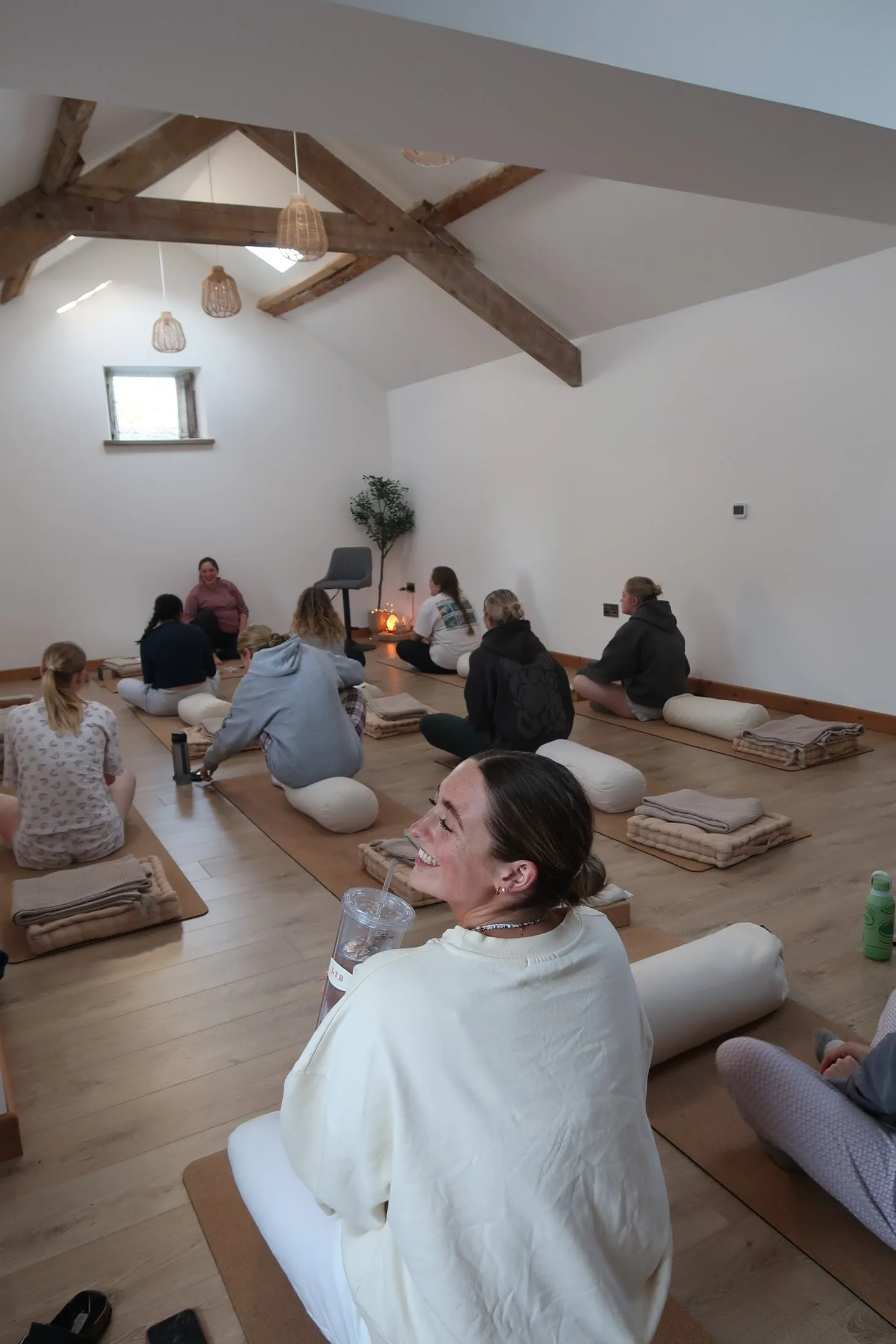 A group on mats facing a woman at the front of a beamed studio, one turning with a big smile