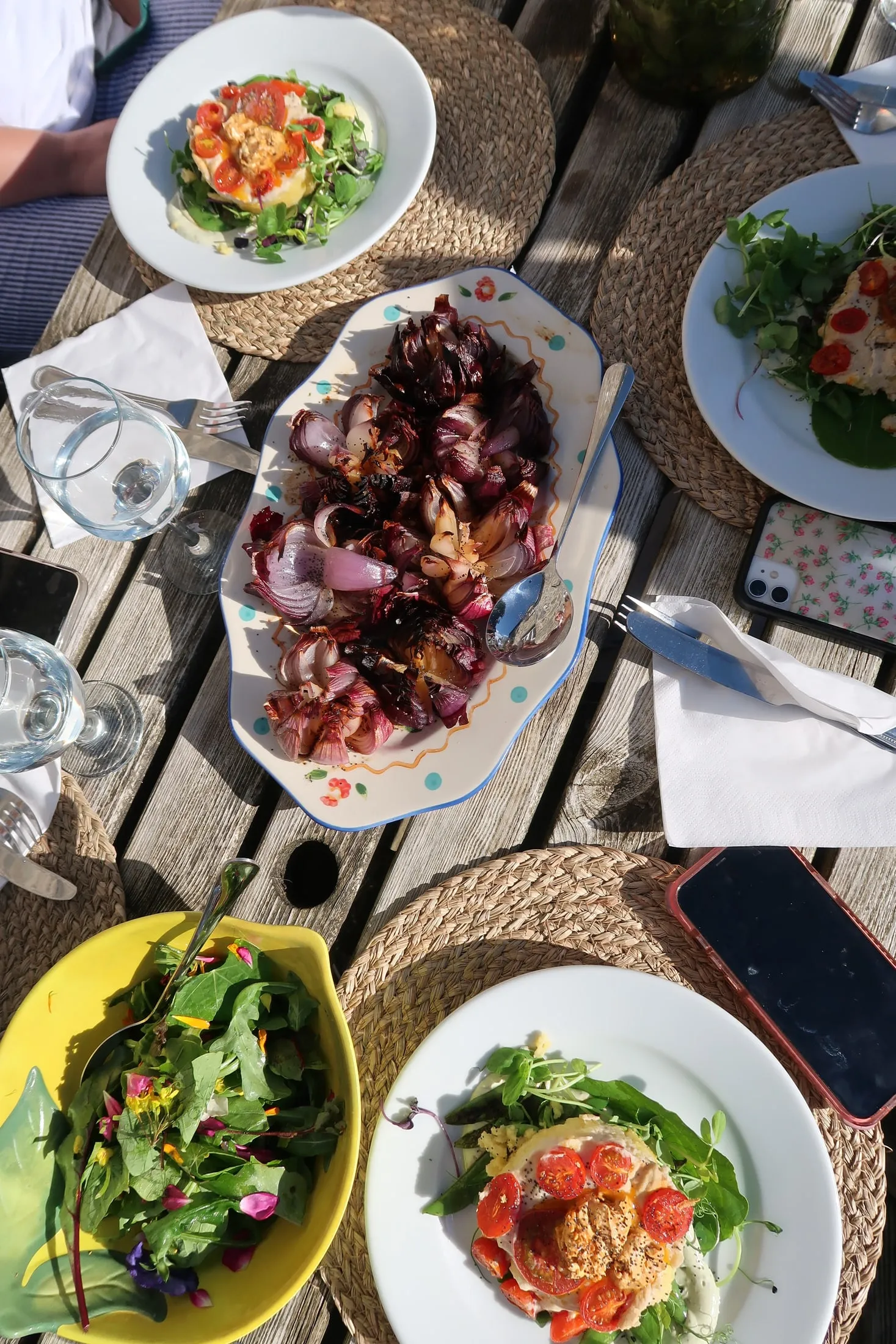 Charred red onions on a painted platter, plates of salad and a bowl of leaves and flowers, seen from above
