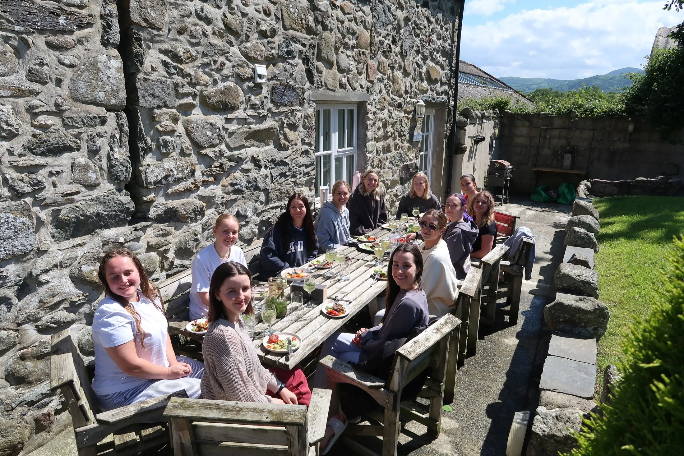 Women smiling round a long outdoor table of salads by a stone house in the sun