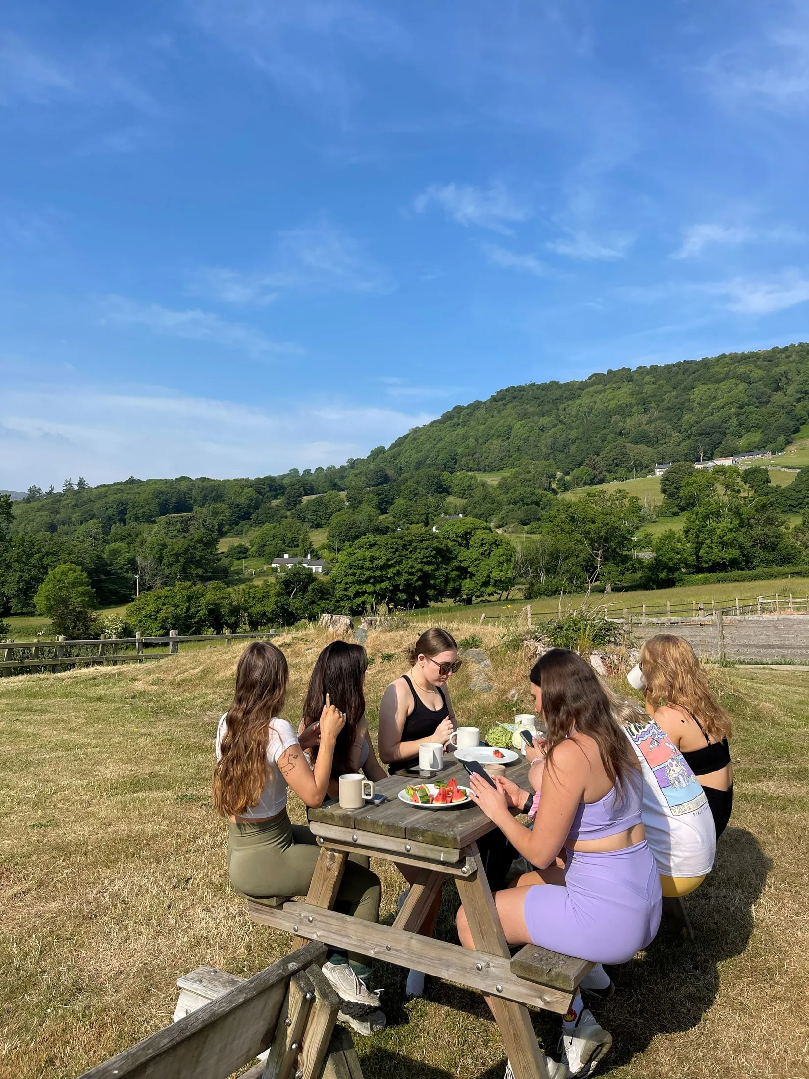 Women eating breakfast at a picnic bench in the sun, hills behind