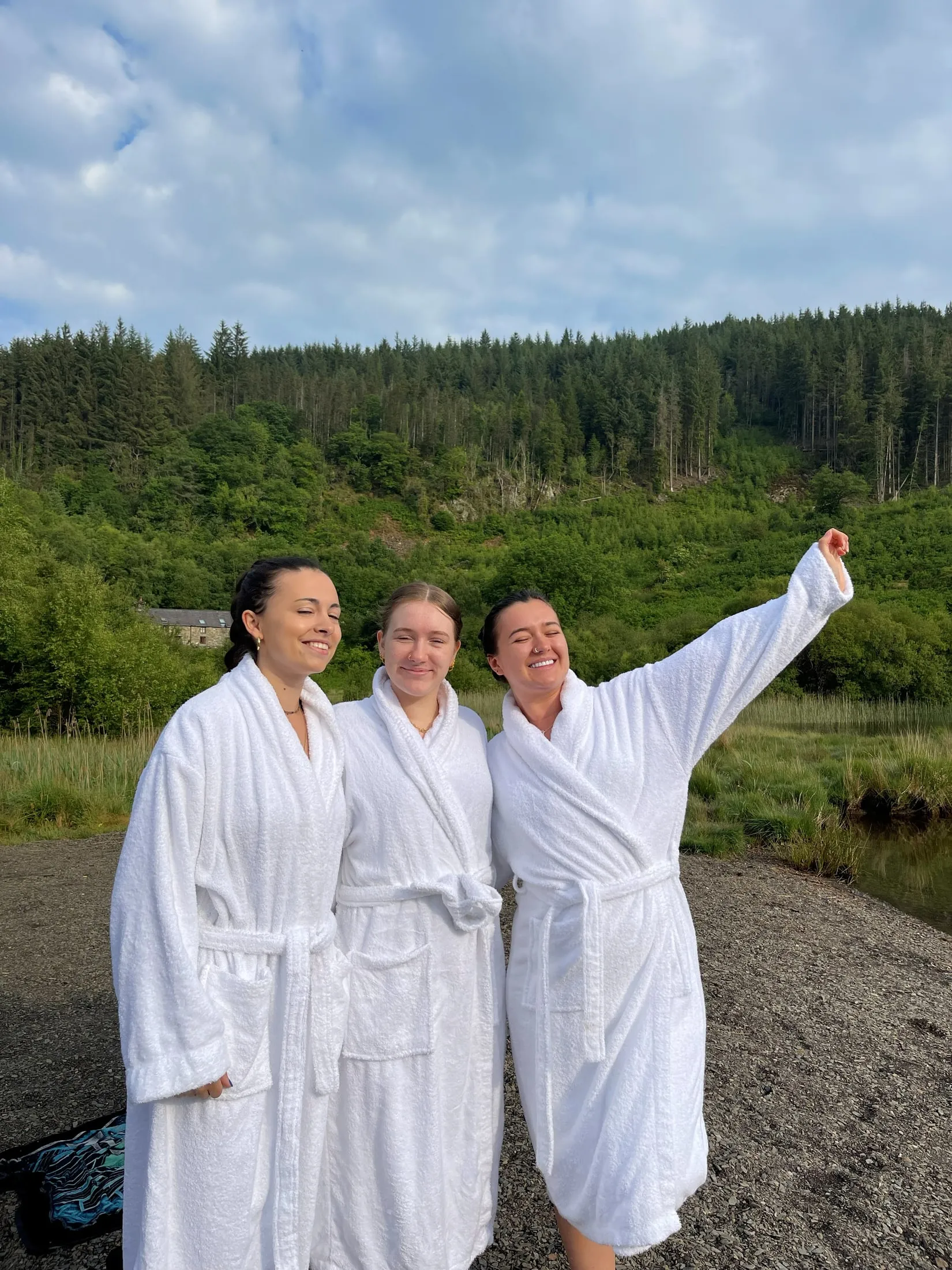 Three women in white dressing gowns by a lake, one with her arm in the air