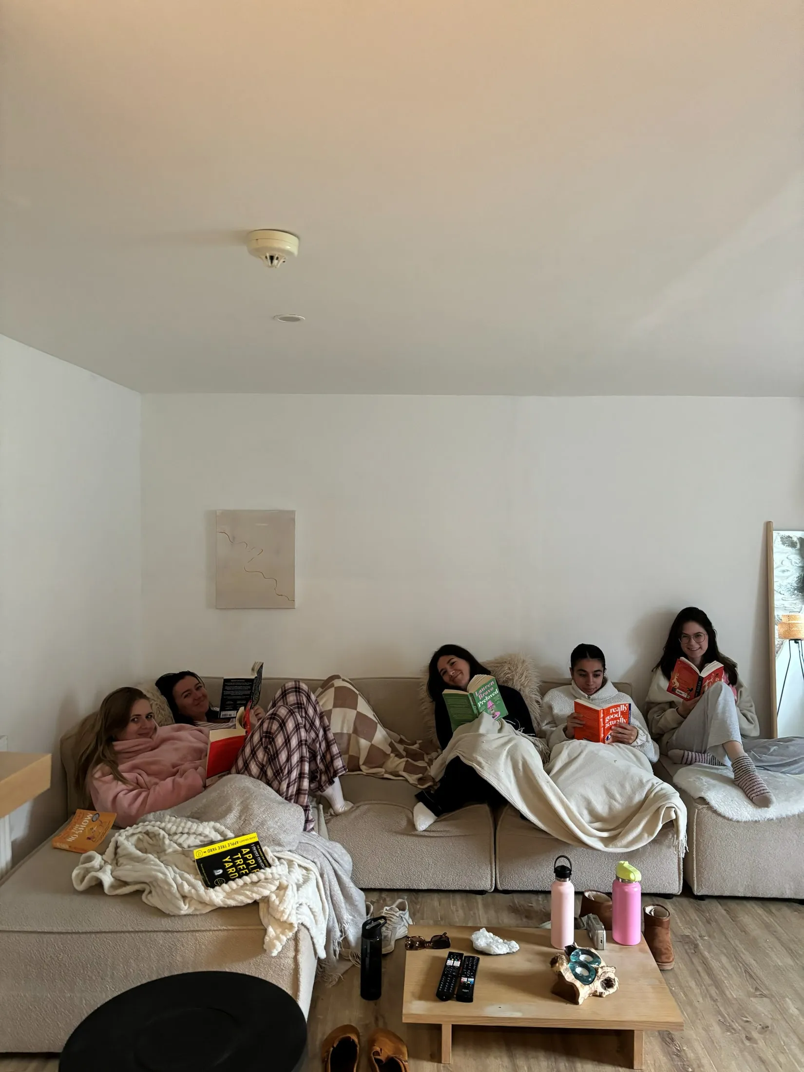 Five women curled up with books and blankets on a big beige sofa