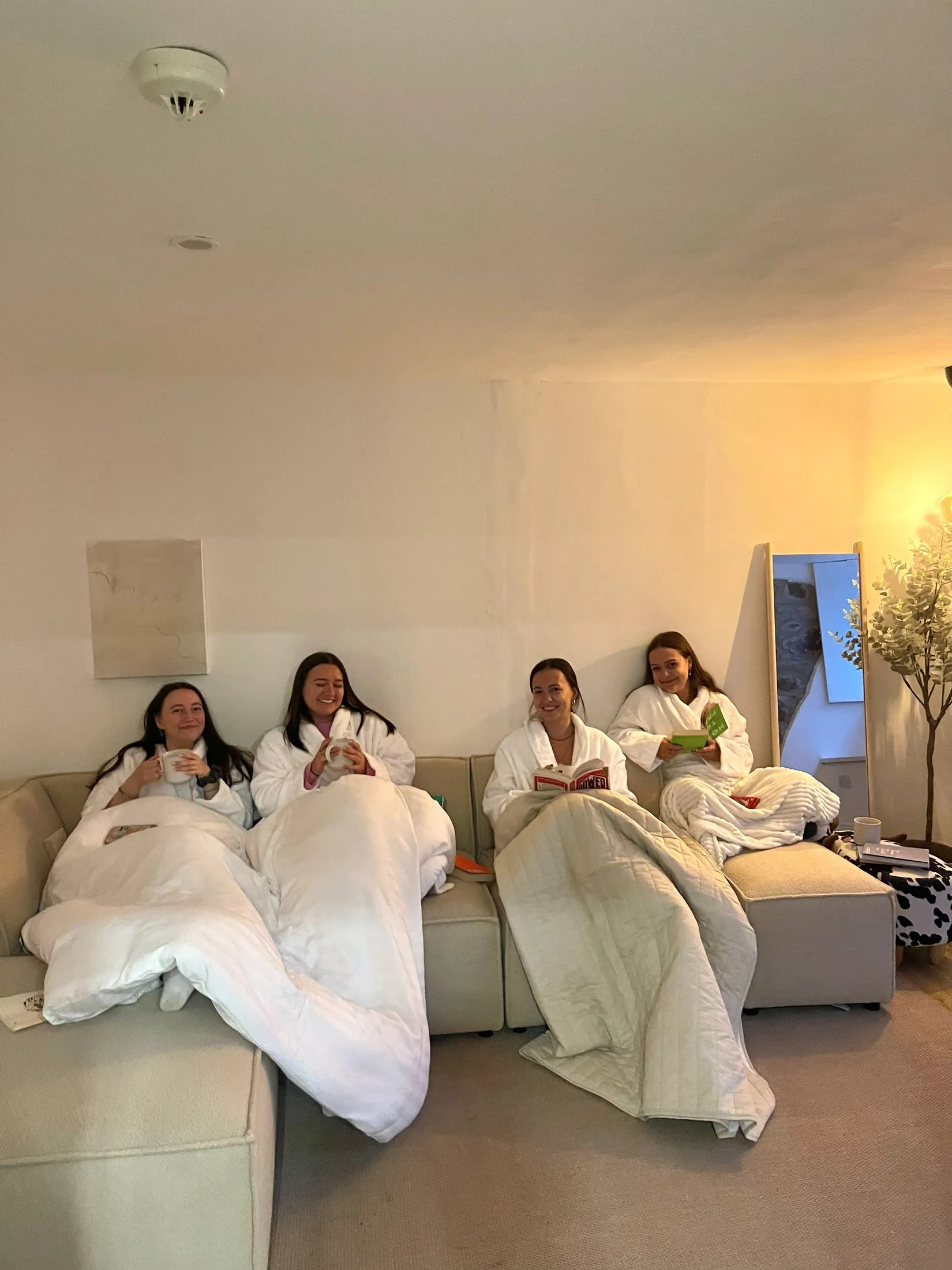 Four women in white dressing gowns tucked under covers on a sofa, with mugs and books