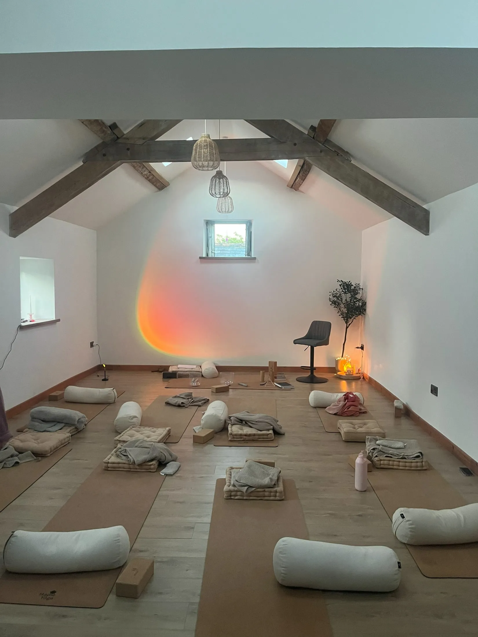 A studio under wooden beams with yoga mats, bolsters and blankets laid out, an orange glow on the wall