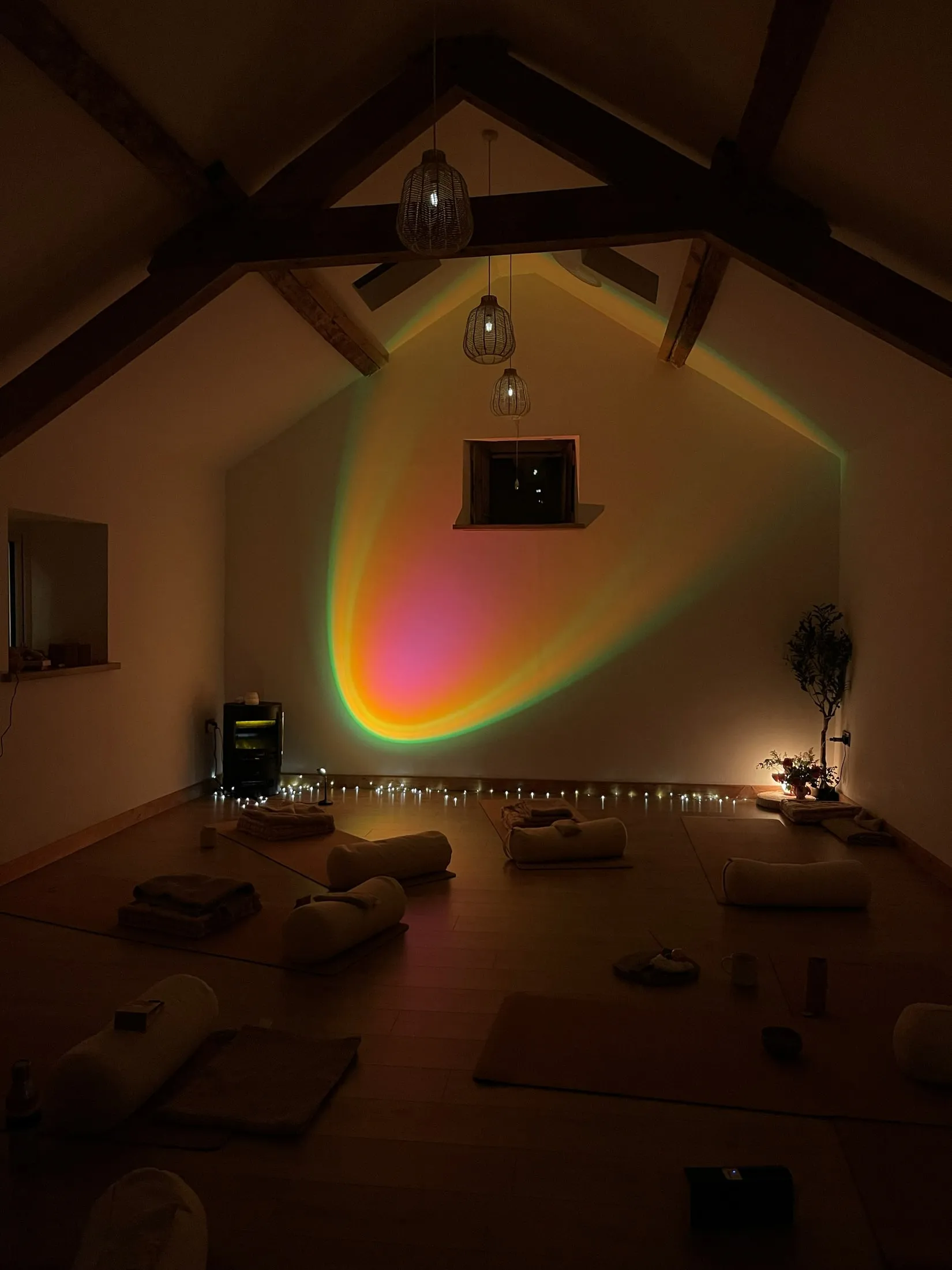 A dark studio lit by a rainbow-edged lamp glow and fairy lights, mats and bolsters laid out