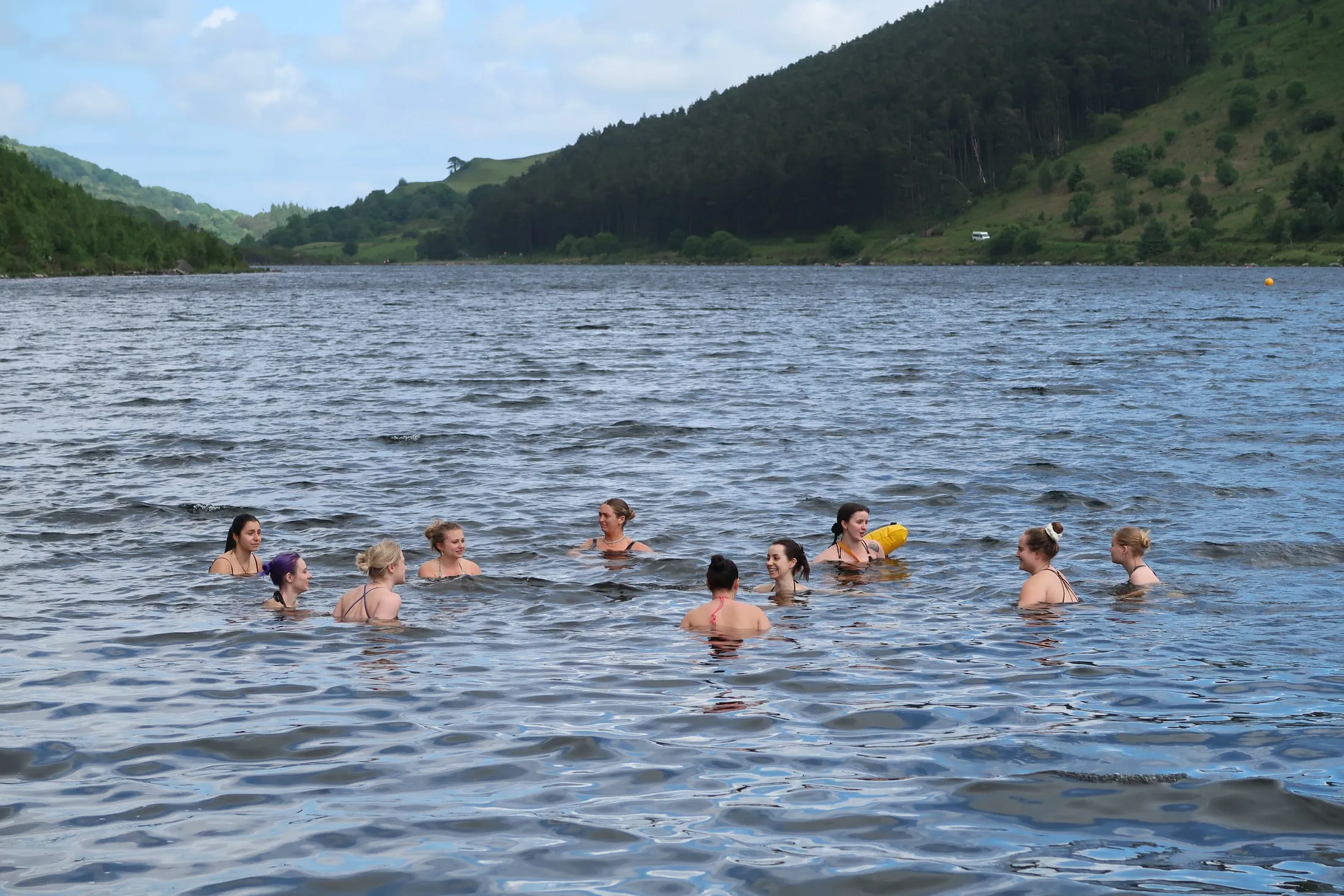 A group of swimmers in a loose circle in a wide lake, forested hills and blue sky behind