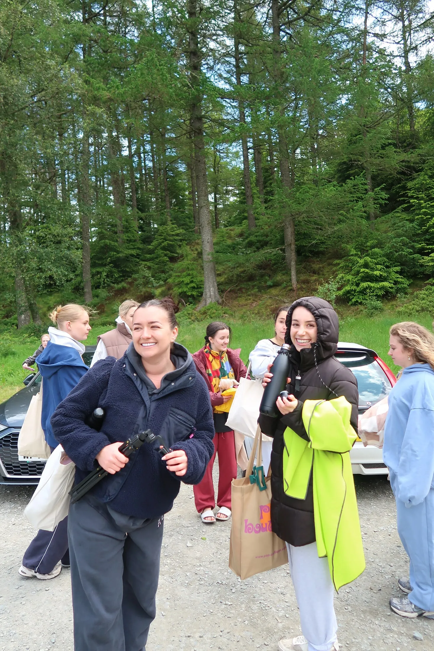 Smiling women with coats and bags gathered in a gravel car park in front of tall trees